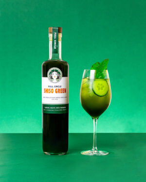 Een fles "Shiso Green" likeur staat naast een groengekleurde cocktail, versierd met komkommerschijfjes en muntblaadjes, tegen een levendige groene achtergrond.
